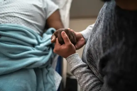 A caretaker holds hands with a hospice patient in a bed in Portland, OR