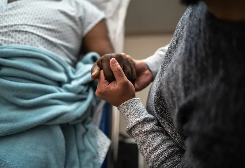 A caretaker holds hands with a hospice patient in a bed in Portland, OR