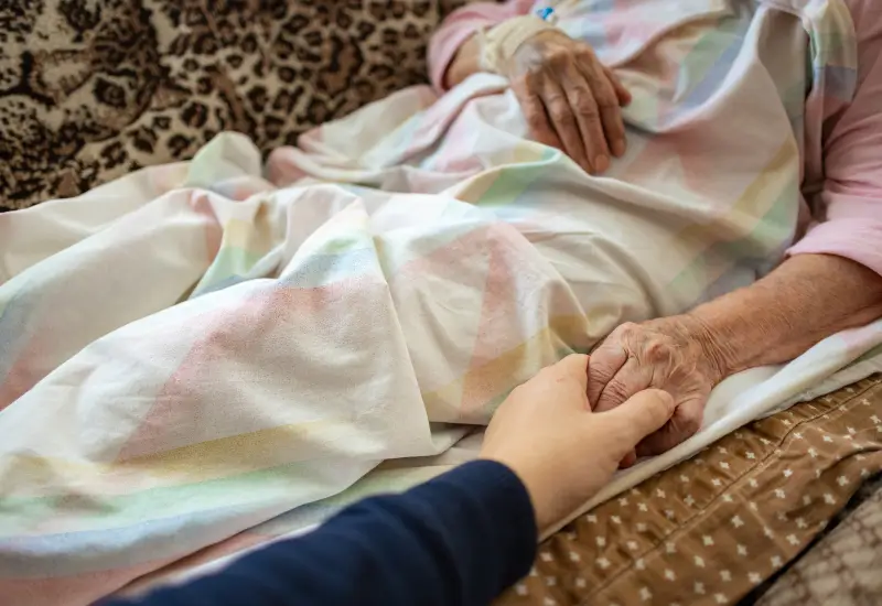 A woman in hospice care lies on her couch and holds hands with someone